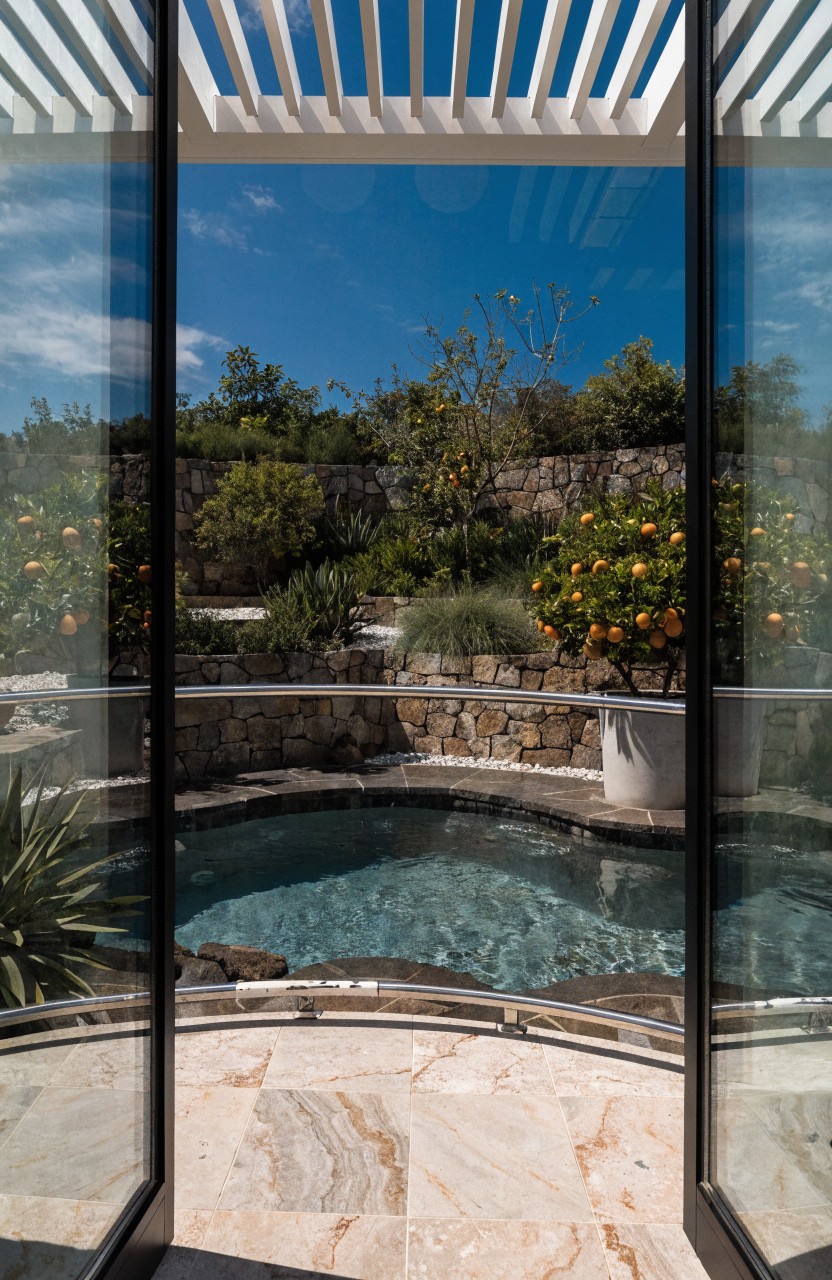 Open glass doors showing a curved blue pool on a stone terrace edged by rock walls, plants, grasses, and orange trees under a clear blue sky.