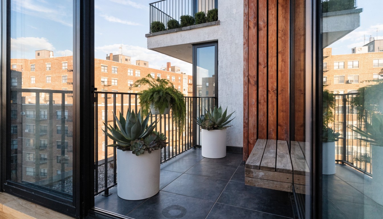 Modern balcony with large textured white pots filled with succulents and ferns, a wooden bench, dark tiled floor, and glass panels reflecting clouds and buildings.