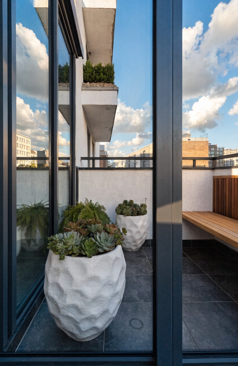 Modern balcony with large textured white pots filled with succulents and ferns, a wooden bench, dark tiled floor, and glass panels reflecting clouds and buildings.