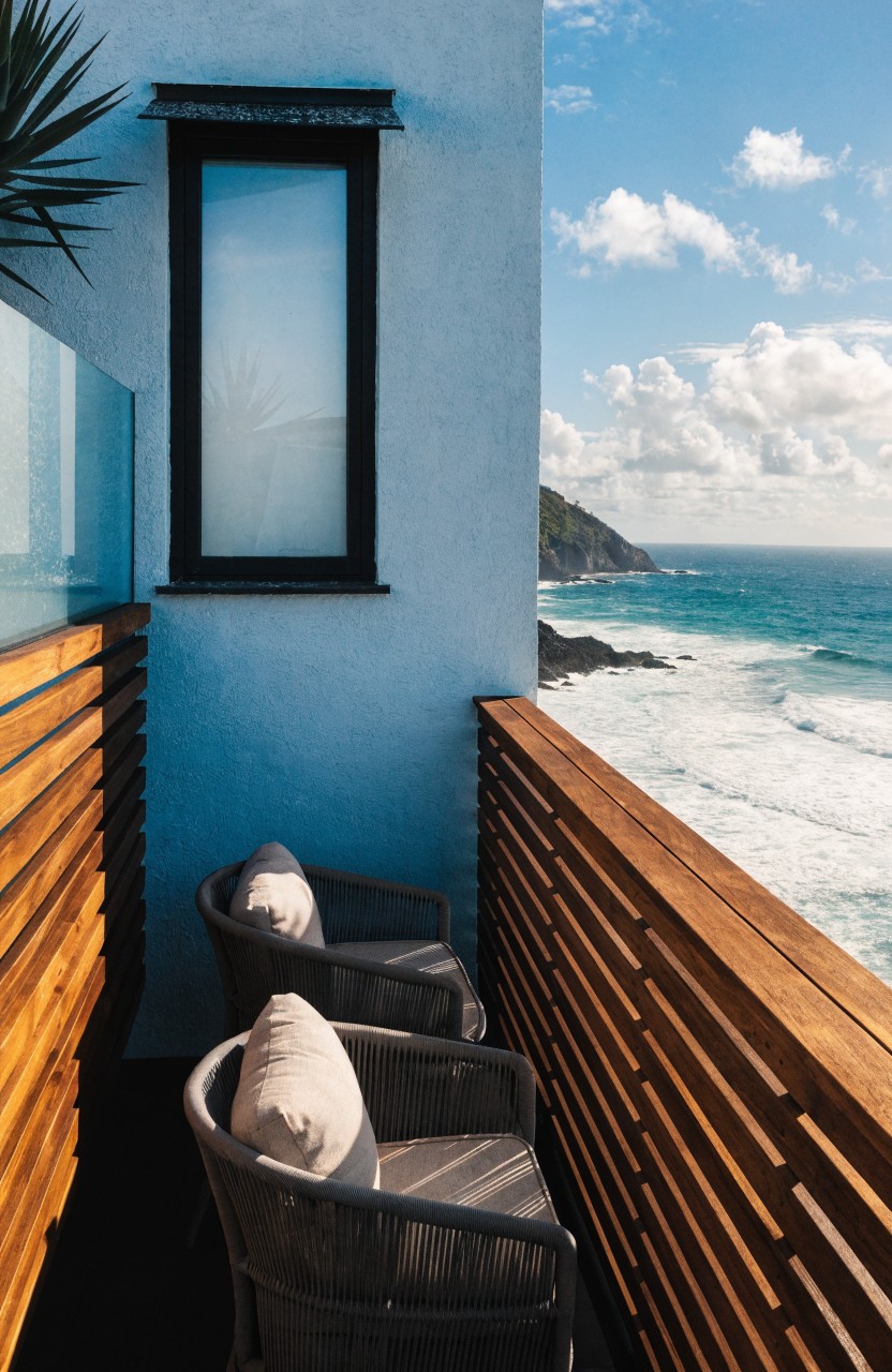 Modern balcony on a blue wall with horizontal wooden slat railings, two gray cushioned chairs, potted palm plants, a frosted window in a black frame, glass panels, and ocean view.
