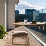 Small outdoor balcony featuring white brick walls, wooden deck flooring, a cantilevered wooden counter with two glasses of iced lime drink, a single cushioned wire bar stool, vertical black planters with greenery, glass railing, and a background view of London skyline with St. Paul's Cathedral.