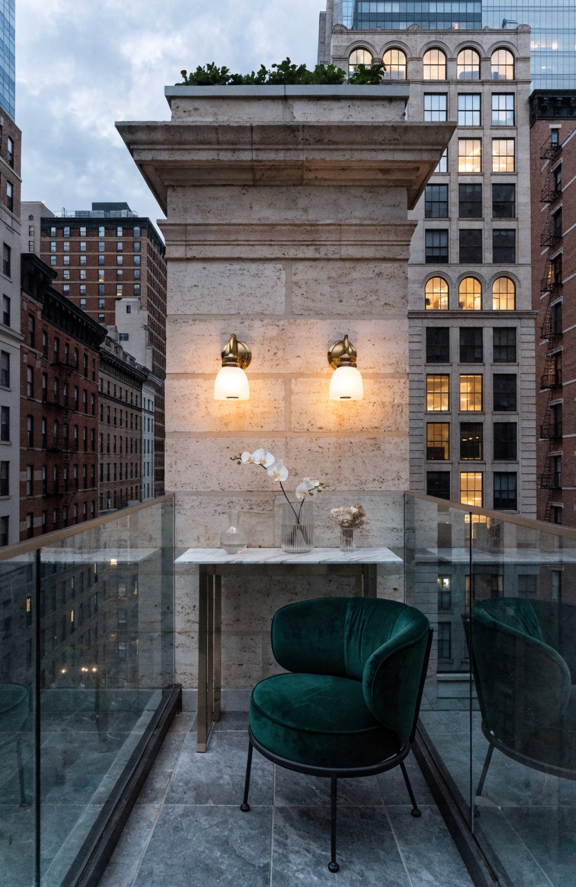 A narrow balcony with gray stone flooring and a tall stone wall holds two curved green velvet armchairs facing a narrow console table topped with white orchids in a vase, lit by two brass wall sconces, behind a glass railing with city buildings in the background.