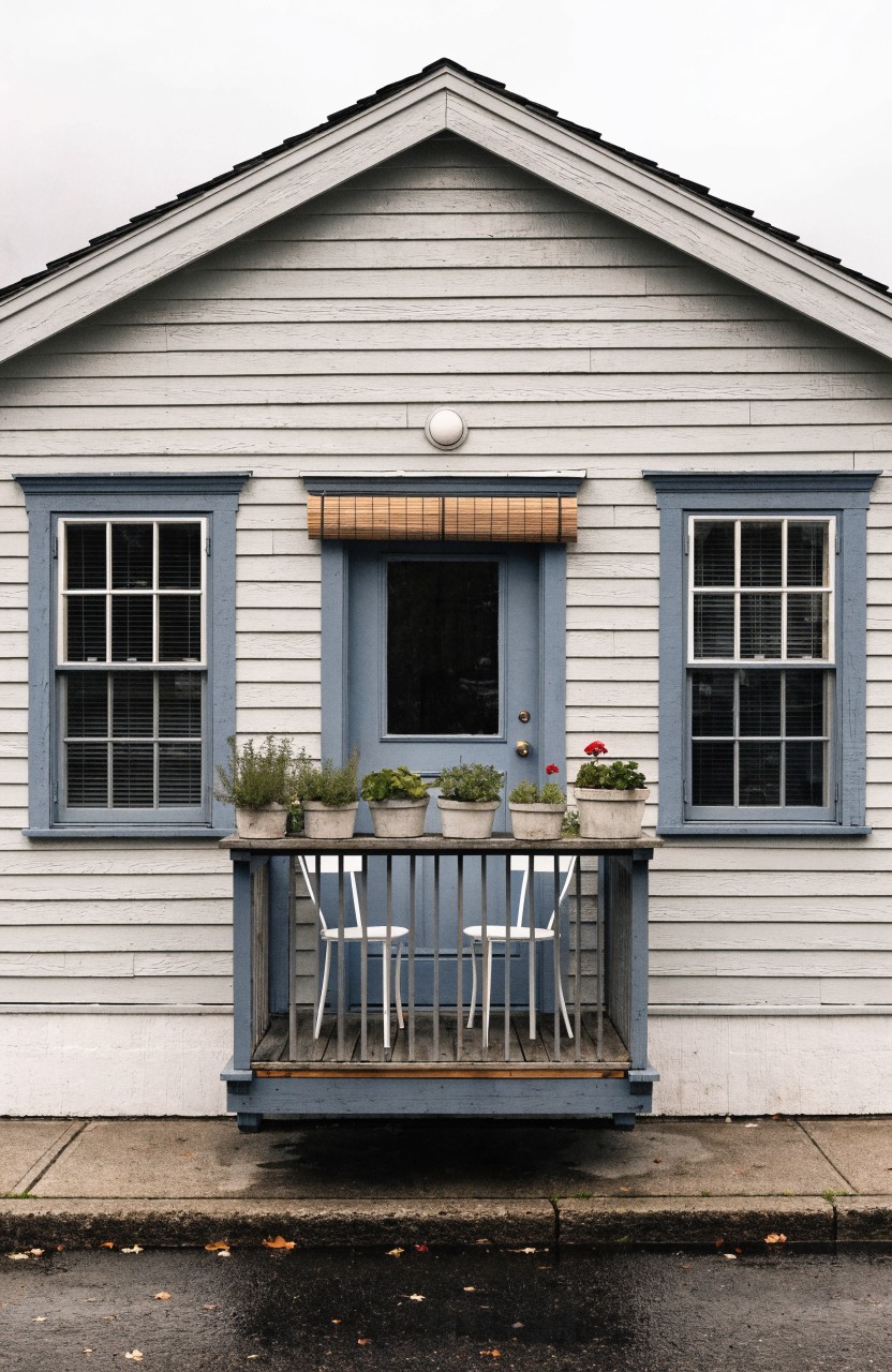 Gray clapboard house with blue window frames and door, small front balcony with white chairs and railing lined with potted plants including herbs and a red flower.