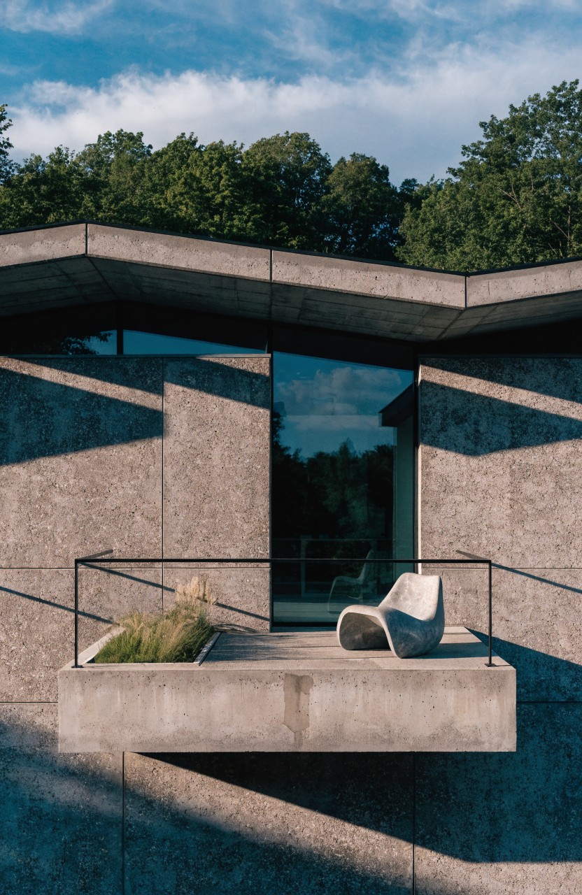 Modern concrete house exterior with a cantilevered balcony holding a white sculptural chair and grass planter next to a large glass window, trees in the background under partly cloudy skies.