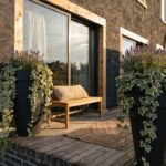 Black metal framed glass enclosure attached to brick house wall, featuring wooden deck floor, cushioned bench, and two large black pots of lavender plants on either side.