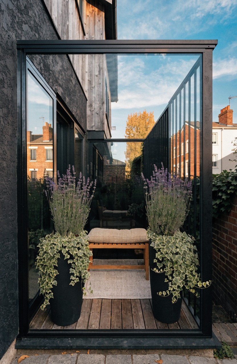 Black metal framed glass enclosure attached to brick house wall, featuring wooden deck floor, cushioned bench, and two large black pots of lavender plants on either side.