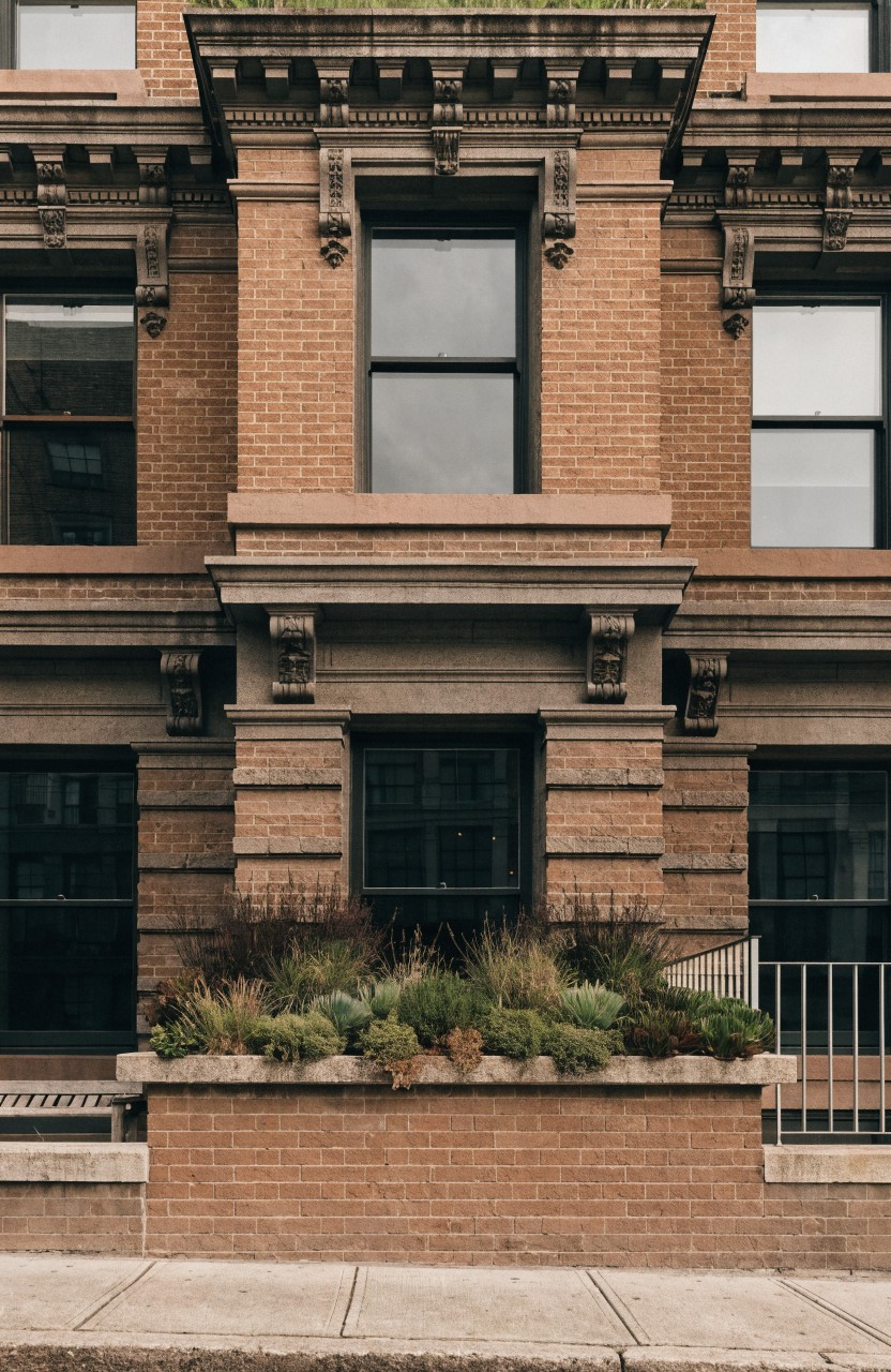 Brick rowhouse facade with large windows, ornate detailing, and a raised planting bed filled with tall grasses along the front sidewalk next to a metal railing.