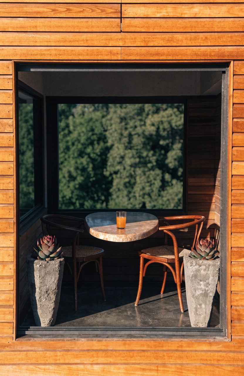 Wood-paneled balcony enclosure with a small round table, two chairs, orange drink glass, and two potted succulents flanking the table, opening via large glass panels to a view of green trees.