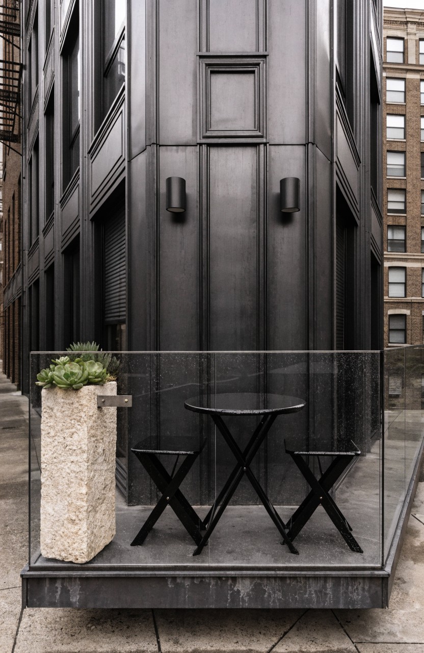 Corner balcony on a dark gray modern building with glass railing, small black X-leg table and two chairs, potted succulent on beige stone pedestal, and wall-mounted lights.