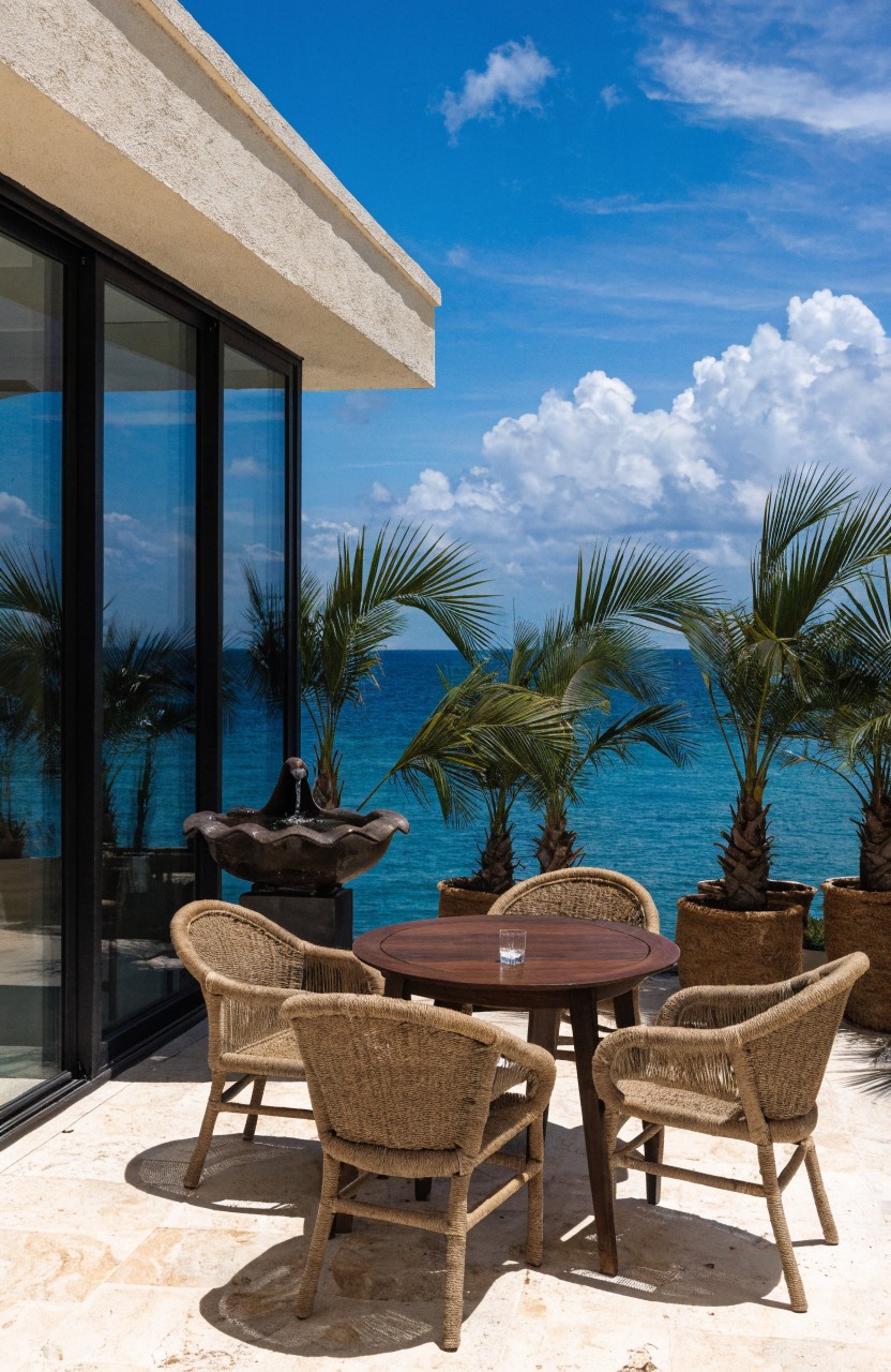 Round wooden table with four rattan armchairs on a light stone terrace balcony next to a beige stucco house wall with black-framed glass doors, surrounded by large potted palms and overlooking a blue ocean.