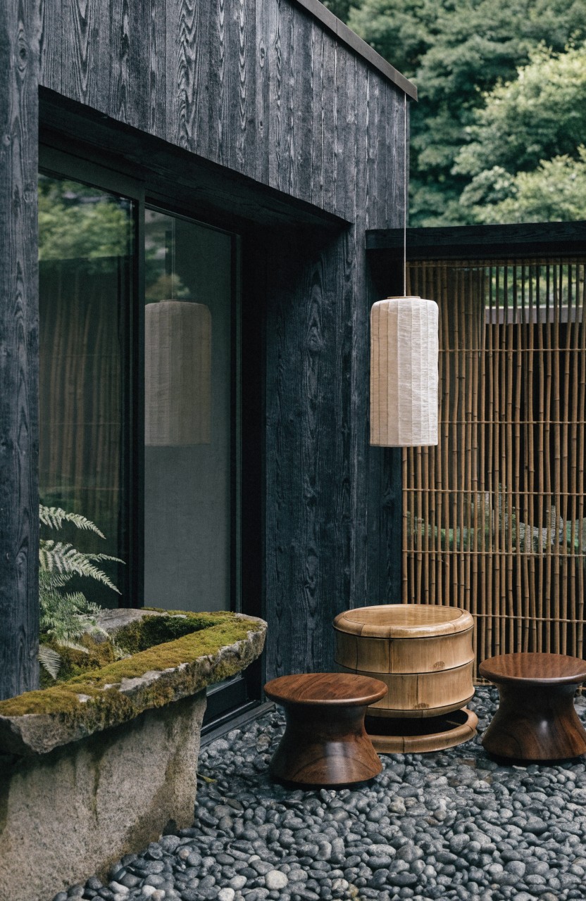 Black charred wood house wall with large sliding glass door opens to gravel patio holding two low walnut stools and one wooden tub stool around mossy stone basin, plus bamboo screen and hanging paper lantern.