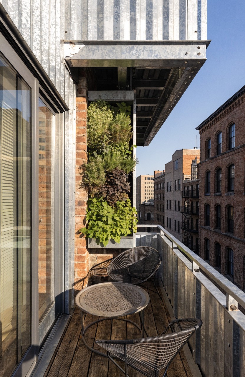 Apartment balcony with corrugated metal siding, a brick wall planted densely with green foliage, wooden deck floor, two black wireframe chairs and matching table, glass door, and city views of brick buildings.