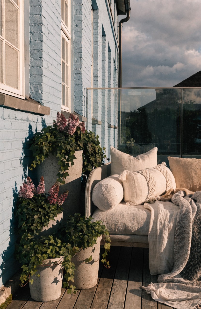 Blue brick wall of a building with a balcony featuring a low white sofa piled with white and beige cushions, pillows, and a gray throw, flanked by two large white pots of purple lilac flowers and trailing ivy plants, glass panel railing, and wooden deck floor.