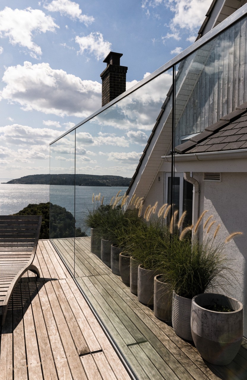 Wooden balcony deck with frameless glass railings lined by tall potted grasses in concrete pots, overlooking the sea next to a white house with pitched roof and chimney.