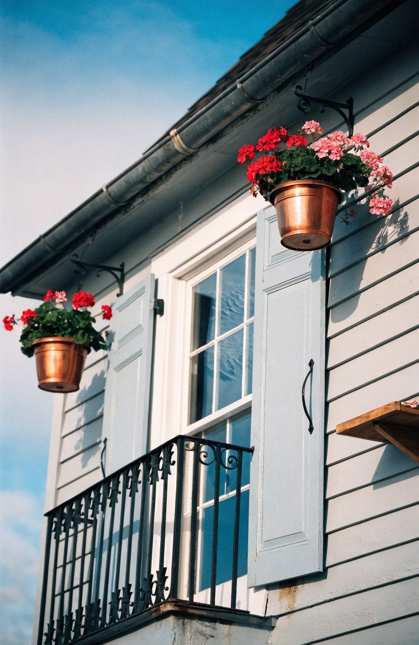 Gray clapboard house exterior with a divided-light window flanked by blue shutters above a black wrought-iron balcony railing, and two copper hanging pots filled with red and pink geraniums mounted on brackets under the roofline.