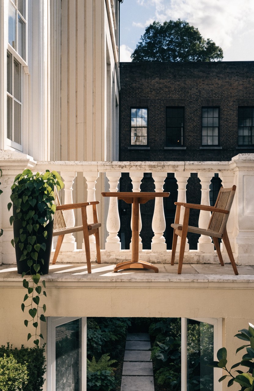 Balcony with two light wooden armchairs and a round pedestal table positioned by a white balustrade, potted green plants nearby, overlooking a stone garden path and brick buildings.