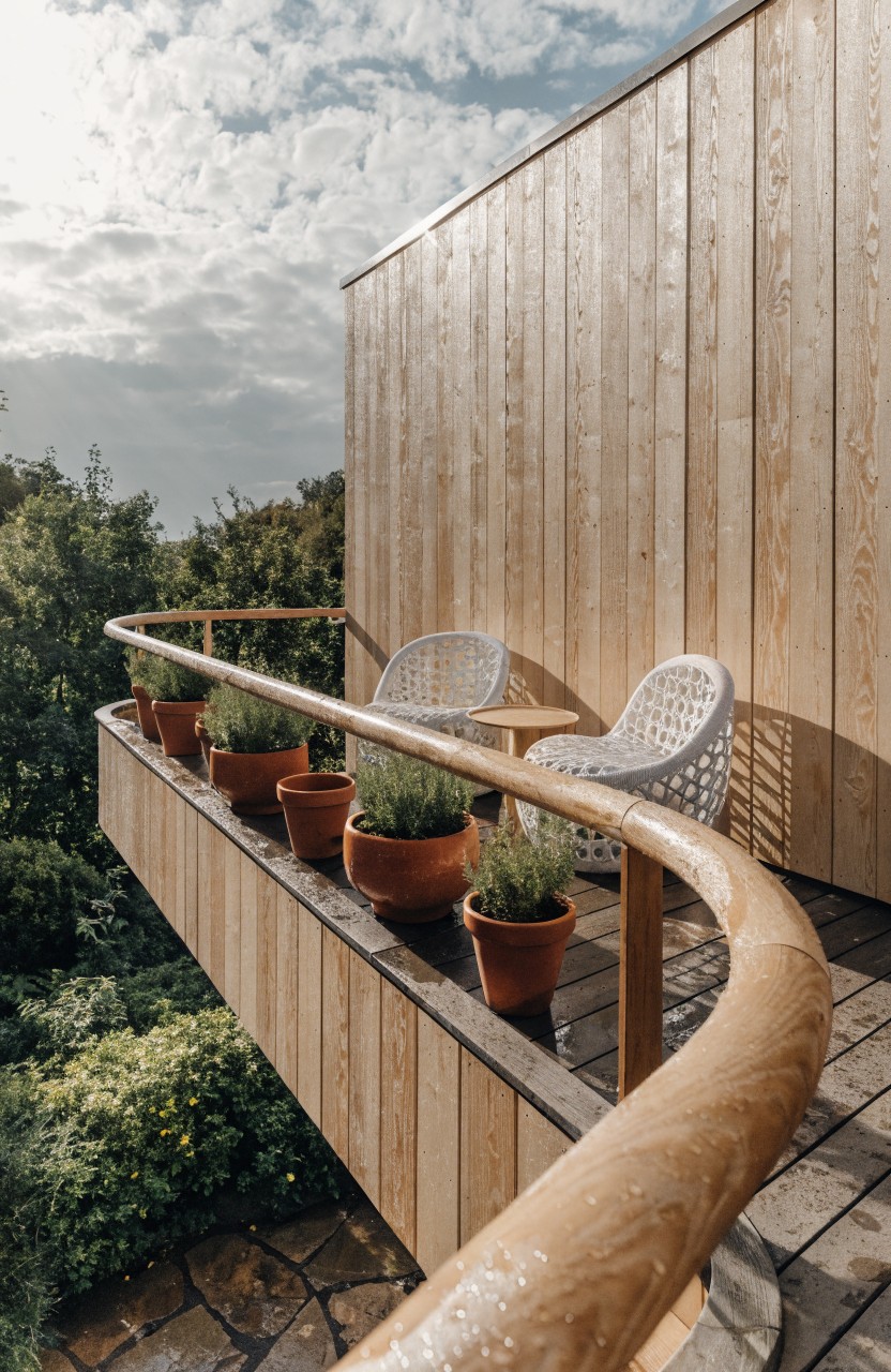 Cantilevered balcony of light wood extending from matching wood-clad house wall, with curved railing holding terracotta pots of plants, two white woven chairs, small round table, and greenery below.