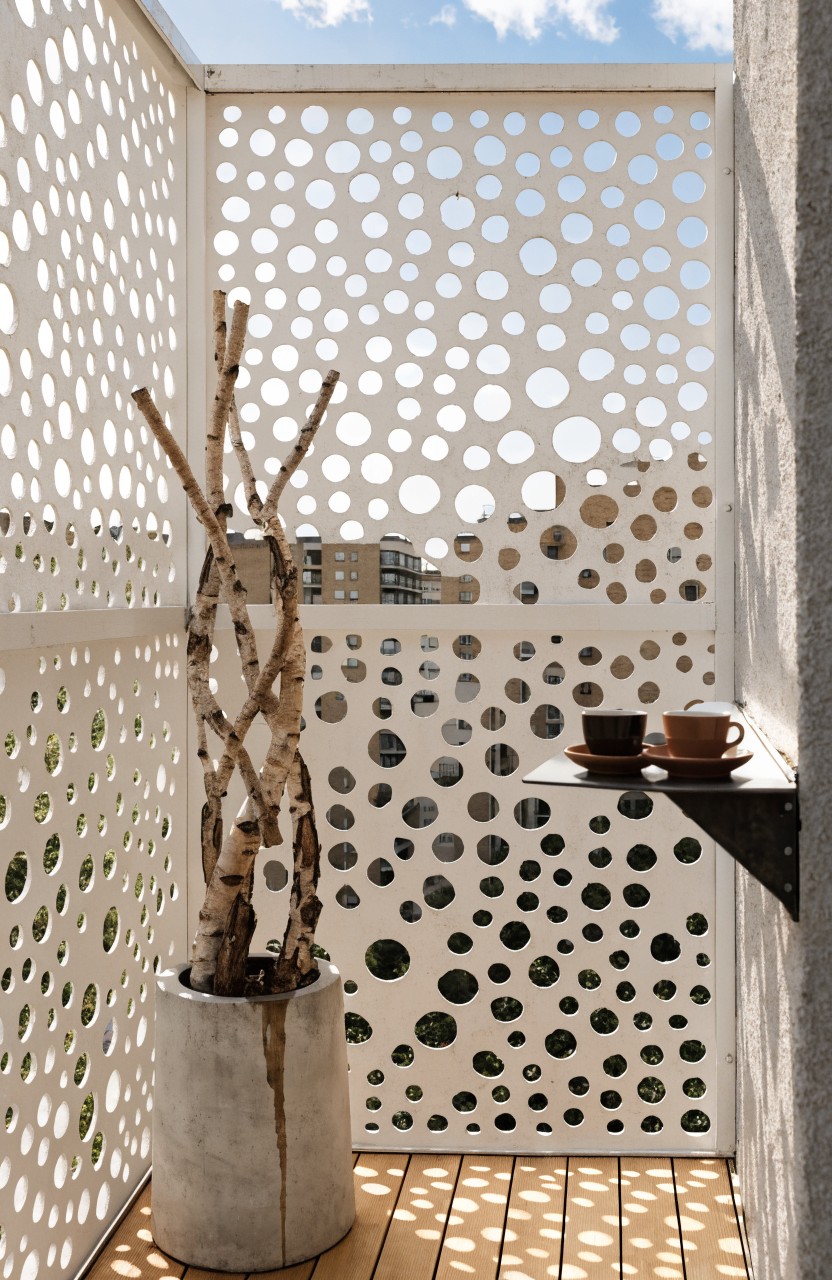White perforated metal screens enclosing a balcony with wooden decking, a potted birch tree with branches, and a shelf holding two teacups, viewed against a cityscape backdrop.