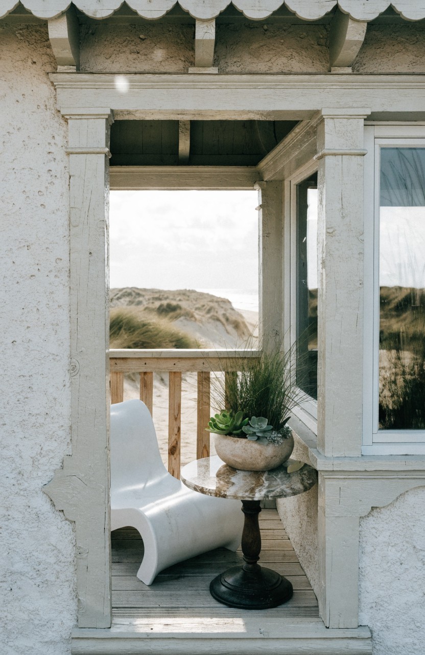 White molded plastic chair and round table with potted succulents and grass on wooden deck balcony framed by white stucco walls and railing, overlooking sand dunes and ocean.