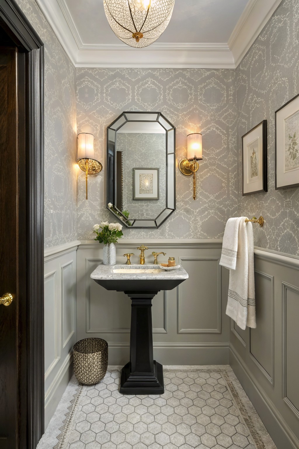 Elegant powder room with soft gray wainscoting below chair rail, gray patterned wallpaper above, black pedestal sink, oval mirror flanked by sconces, and brass accents