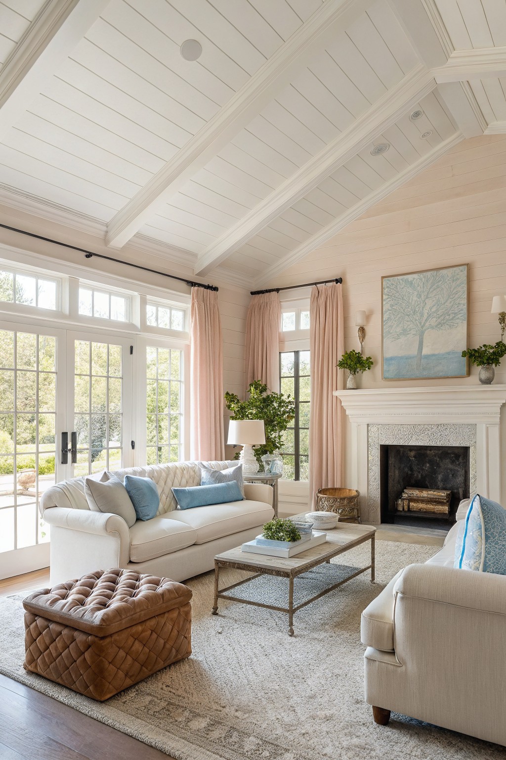 Cozy living room with pale blush shiplap walls, white trim and ceiling beams, cream sofas, tufted leather ottoman, stone fireplace, and large windows to the garden