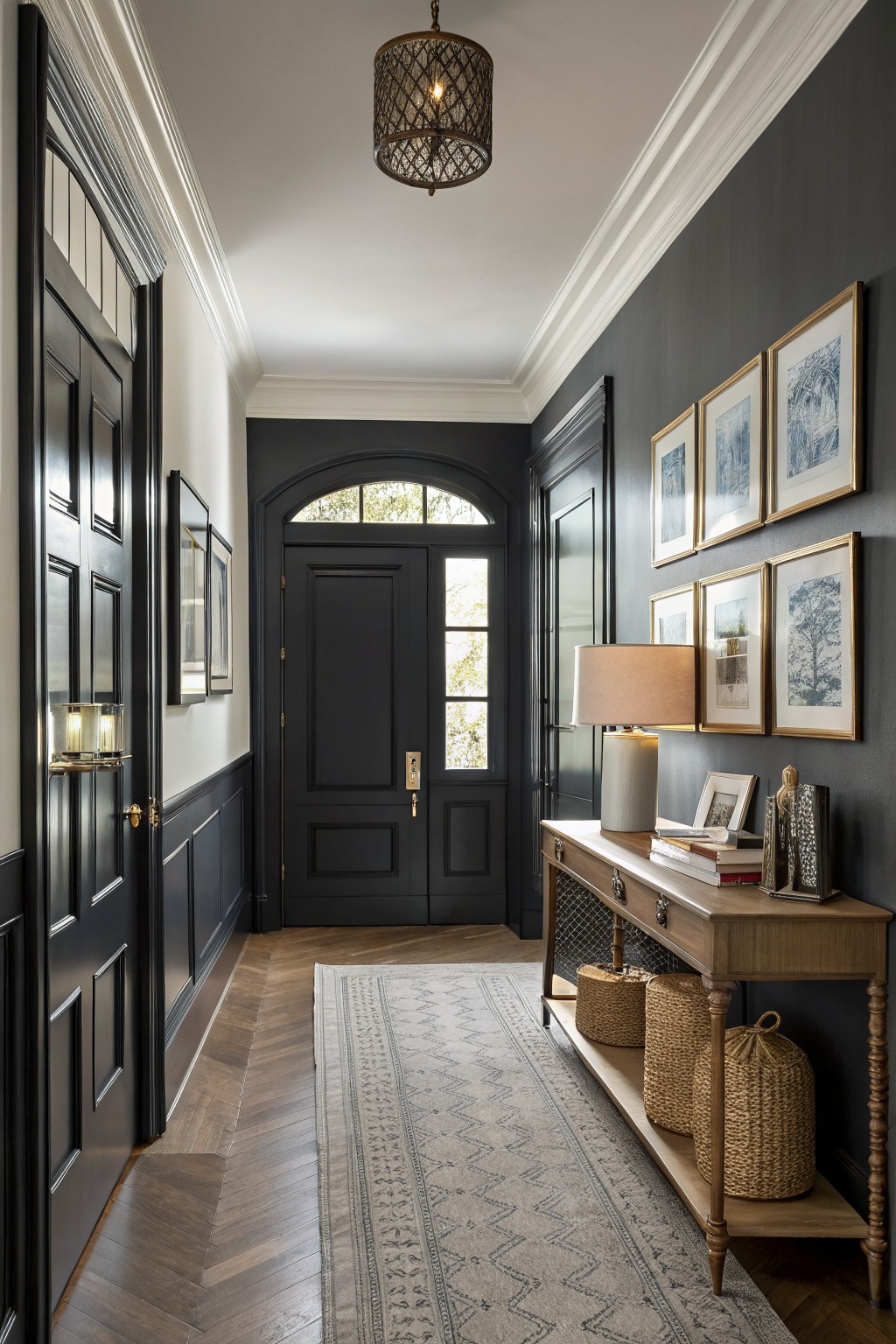 Moody entry hallway with deep charcoal gray walls, wainscoting, arched black door, herringbone wood floors, console table with lamp and baskets, and framed art.