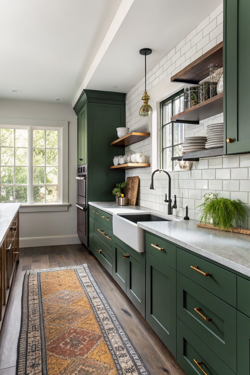 Kitchen with deep green lower cabinets, white subway tile backsplash, farmhouse sink, brass hardware, wood floating shelves, and a window letting in light