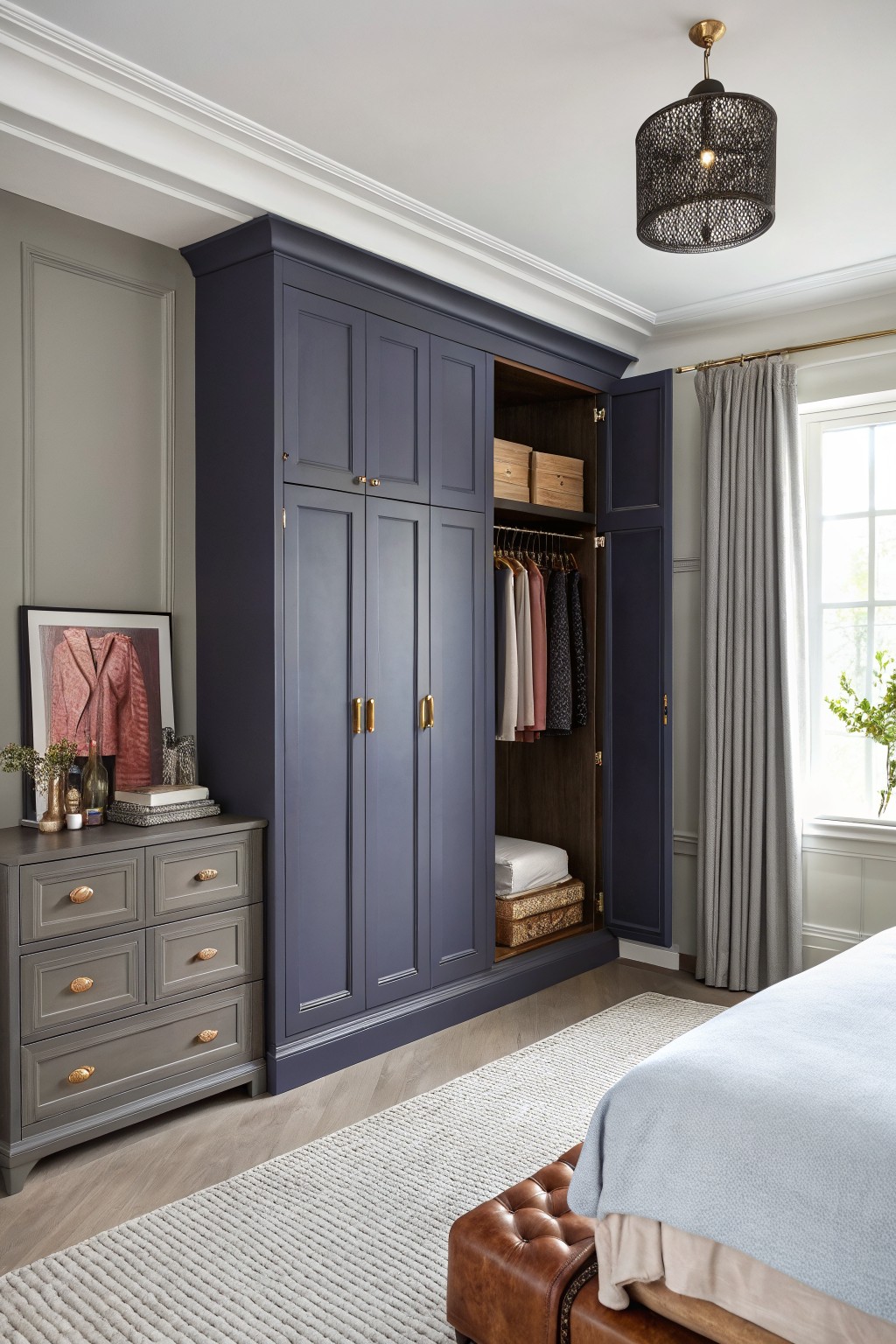 Fitted navy blue wardrobe with open doors showing hanging clothes and shelves, beside a gray dresser in a light gray bedroom with wood floors and window curtains