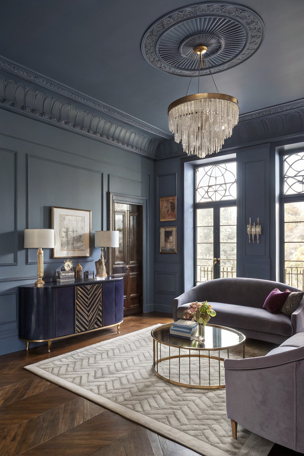Living room with deep navy paneled walls, purple velvet sofa, gold chandelier, navy credenza, and tall windows overlooking balcony