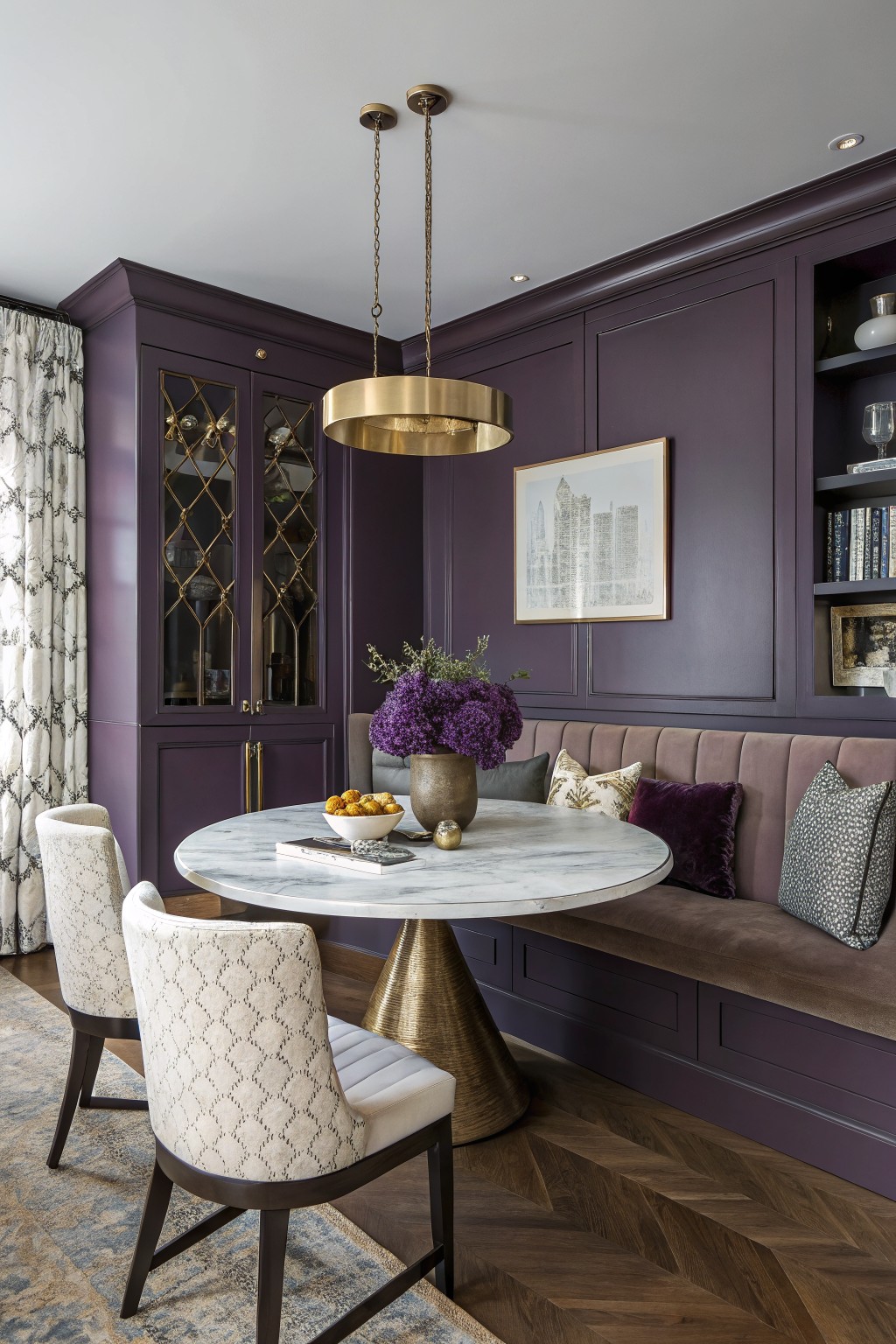 Dining nook with deep purple paneled walls and cabinetry, gold chandelier, marble pedestal table, tufted banquette, and upholstered chairs on herringbone wood floors