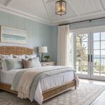 Bedroom with soft blue-gray shiplap walls, rattan bed, white bedding, wood nightstands, and large windows overlooking water