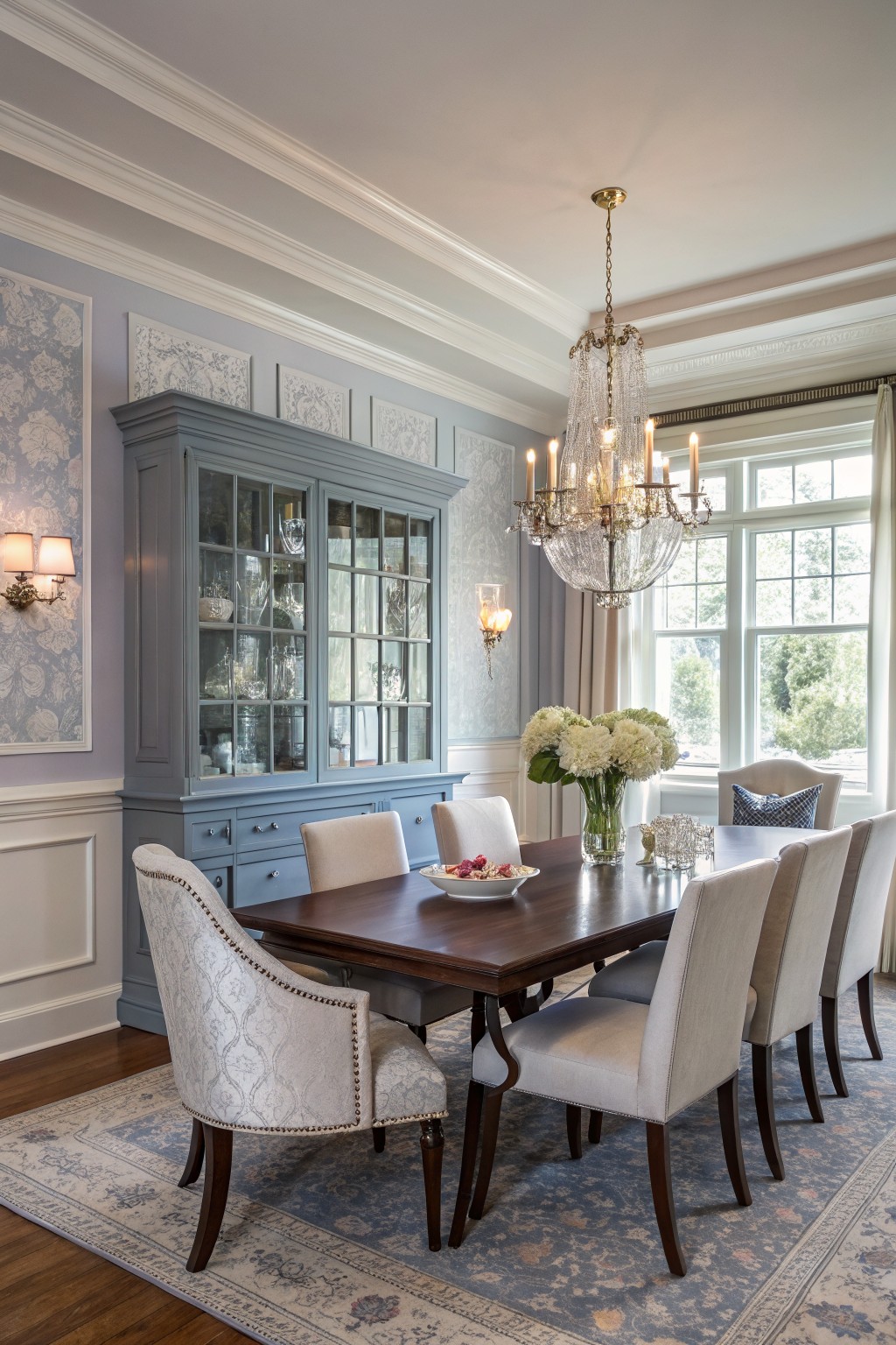 Dining room with pale blue-gray walls, blue-gray china cabinet filled with dishes, wooden table with white upholstered chairs, crystal chandelier, and large sunny windows