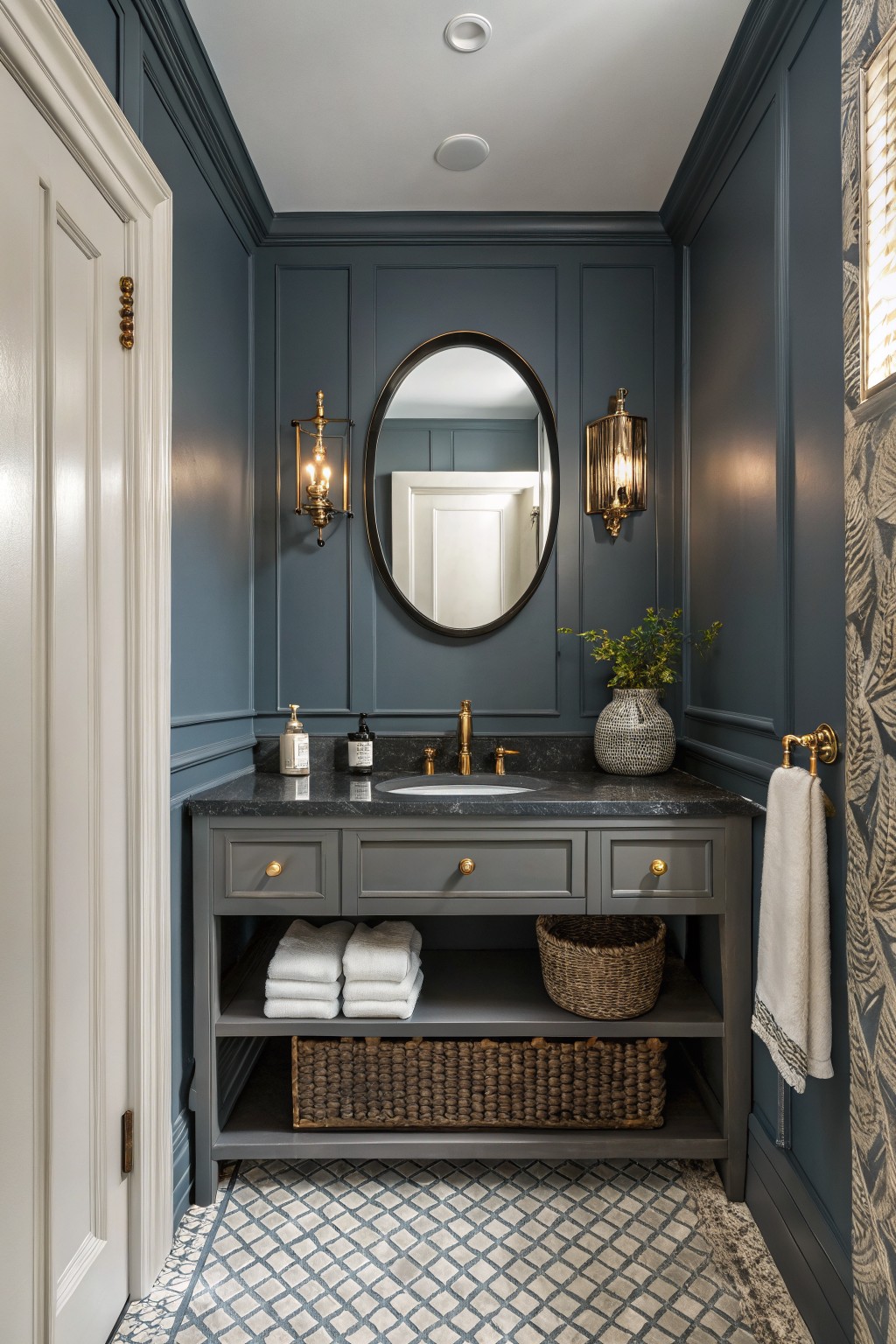 Cozy powder room featuring deep navy paneled walls, oval black-framed mirror with brass sconces, gray vanity with black granite top, and blue-gray patterned tile floor