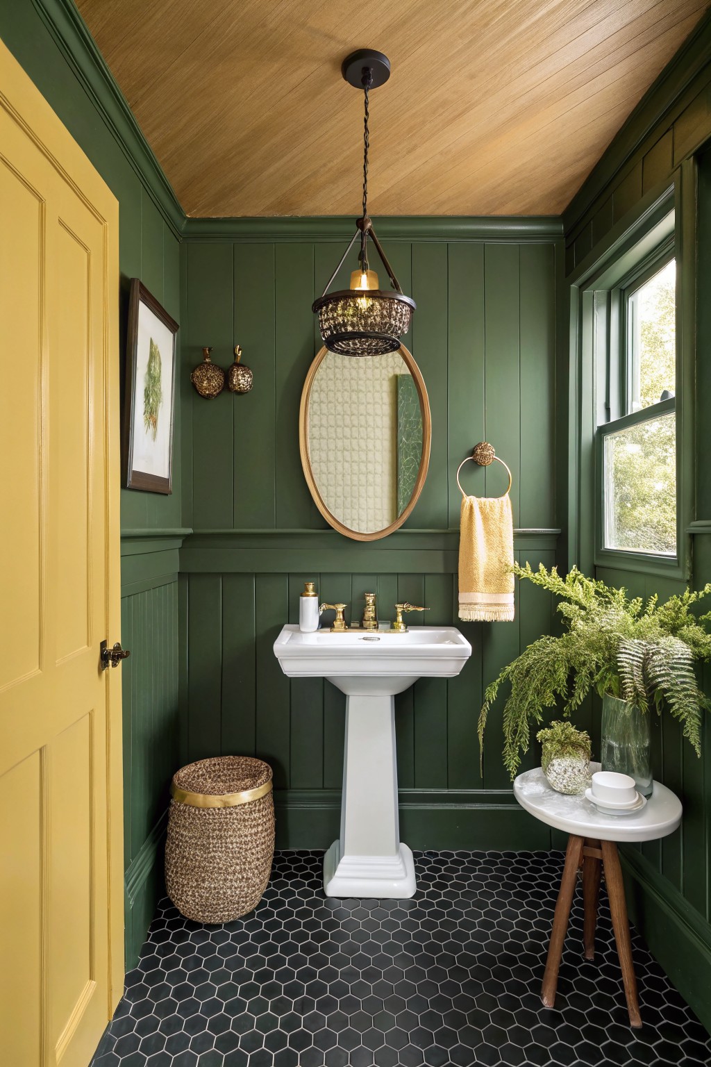 Cozy small bathroom with deep green shiplap walls, yellow door, white pedestal sink, brass fixtures, round mirror, yellow towel, plants, black hexagonal tile floor, and warm wood ceiling