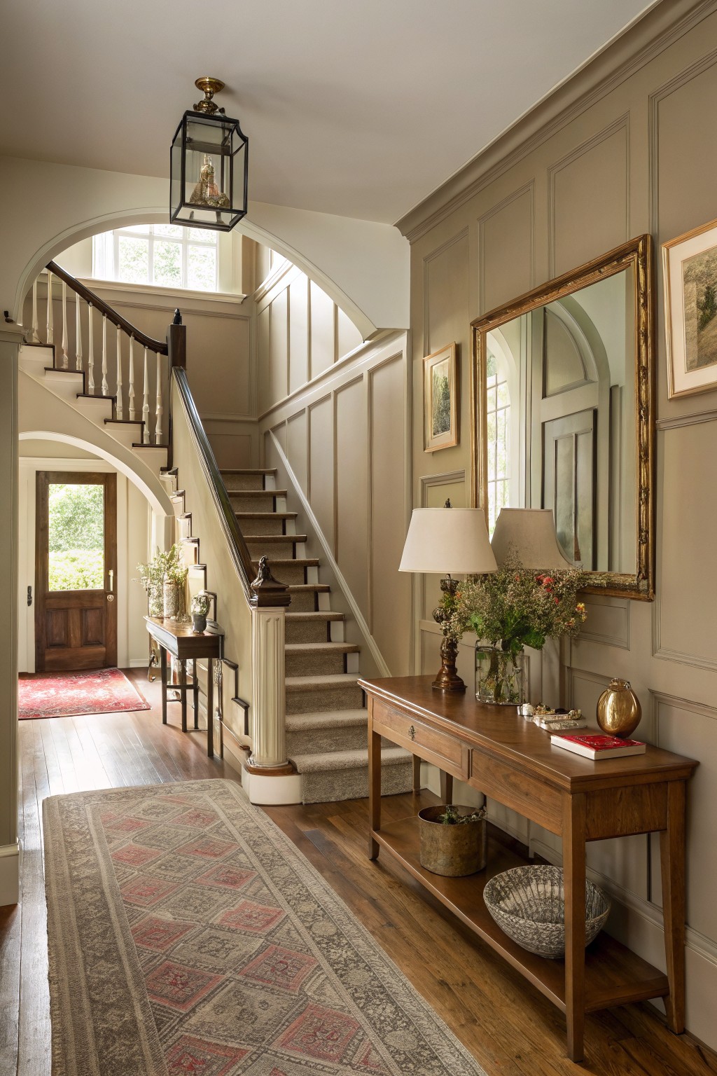 Elegant entry hall with warm greige paneled walls, sweeping staircase, wood console table, and gilded mirror