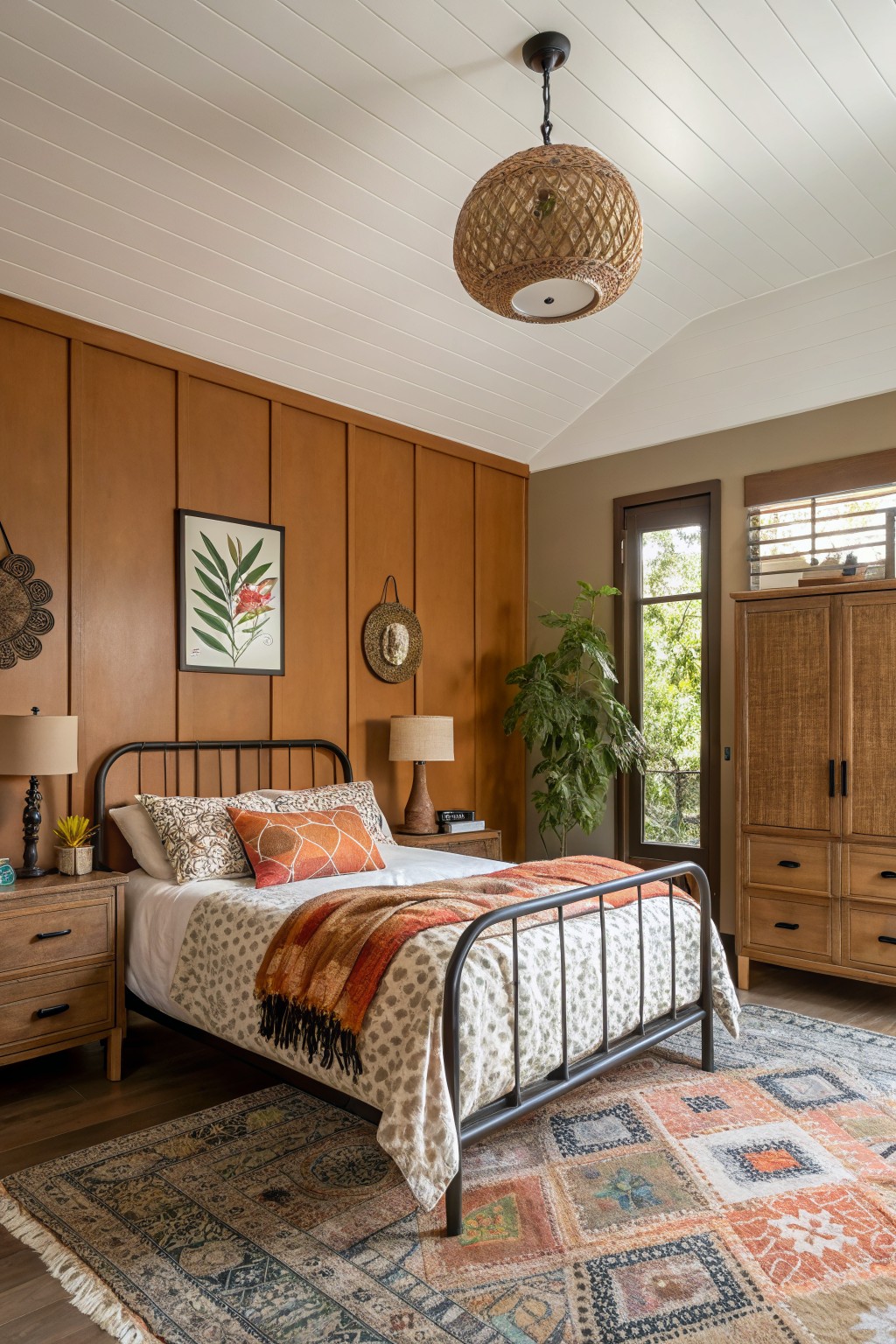 Bedroom with warm brown wood-paneled accent wall, black metal bed frame with orange and patterned bedding, woven pendant light, potted plants, and rattan armoire beside a large window