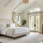 Serene bedroom with warm off-white walls, beige upholstered bed, wood nightstands, layered pillows, and French doors with cream drapes overlooking trees