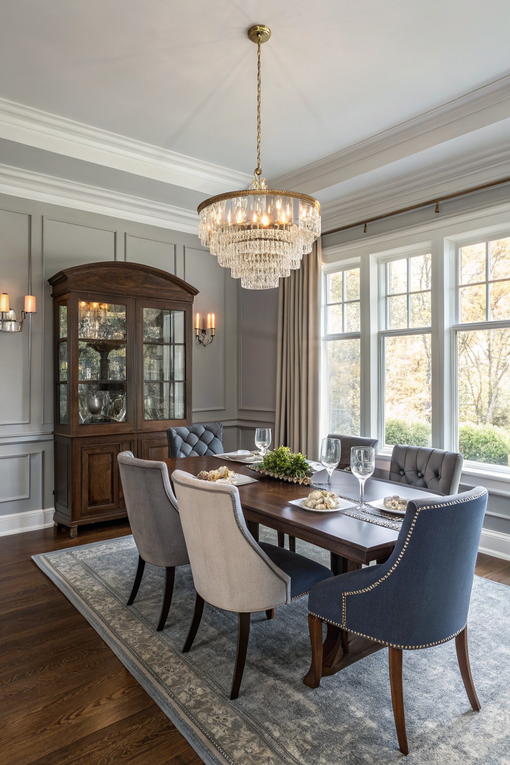Elegant dining room featuring soft gray walls, a sparkling crystal chandelier, warm wood furniture including a china cabinet and table, tufted chairs in gray and blue, and large windows with fall views.
