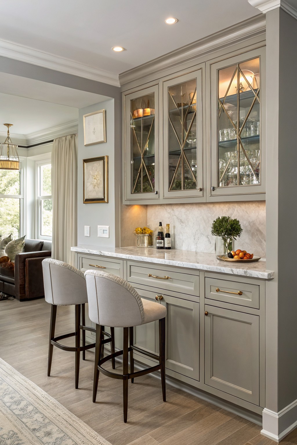 Closeup of elegant kitchen bar with soft greige cabinets, gold accents, marble counters, bar stools, and greenery