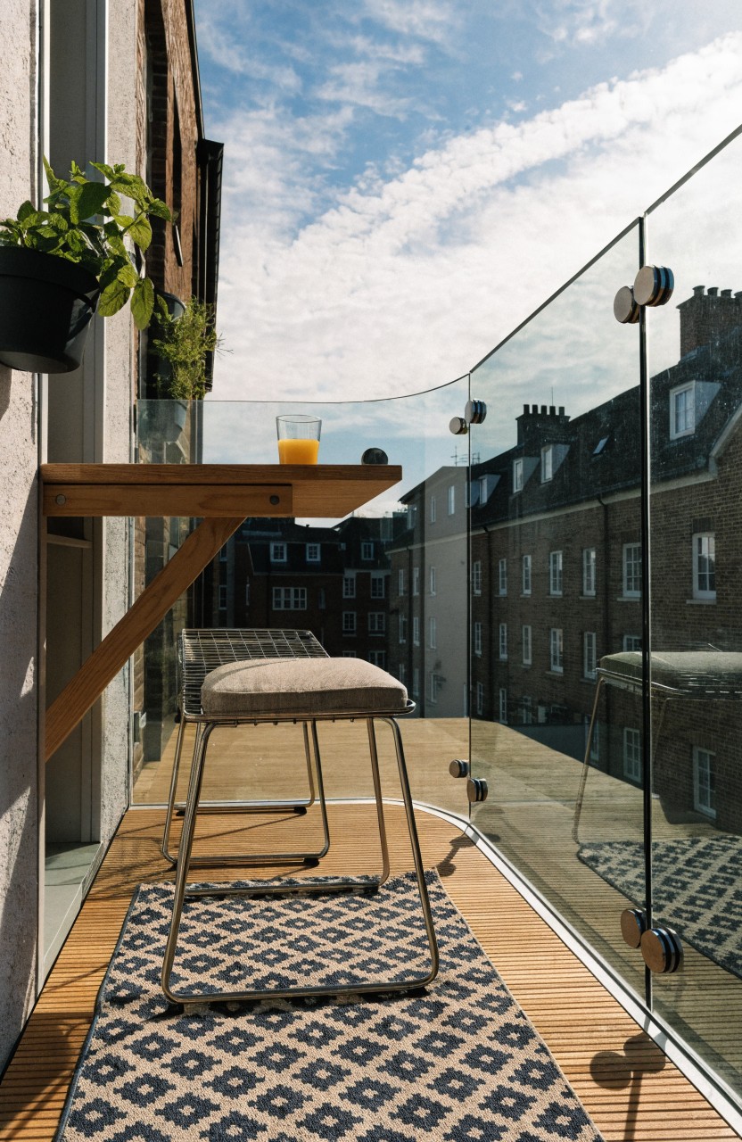 Small outdoor balcony featuring white brick walls, wooden deck flooring, a cantilevered wooden counter with two glasses of iced lime drink, a single cushioned wire bar stool, vertical black planters with greenery, glass railing, and a background view of London skyline with St. Paul's Cathedral.