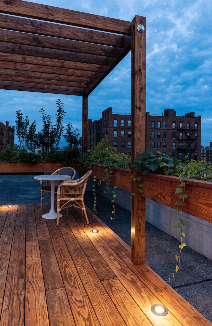 Small rooftop balcony at dusk with wooden pergola, round metal table holding wine glasses and grapes, two curved chairs, linear planters along brick-edged railing, and city buildings visible beyond.