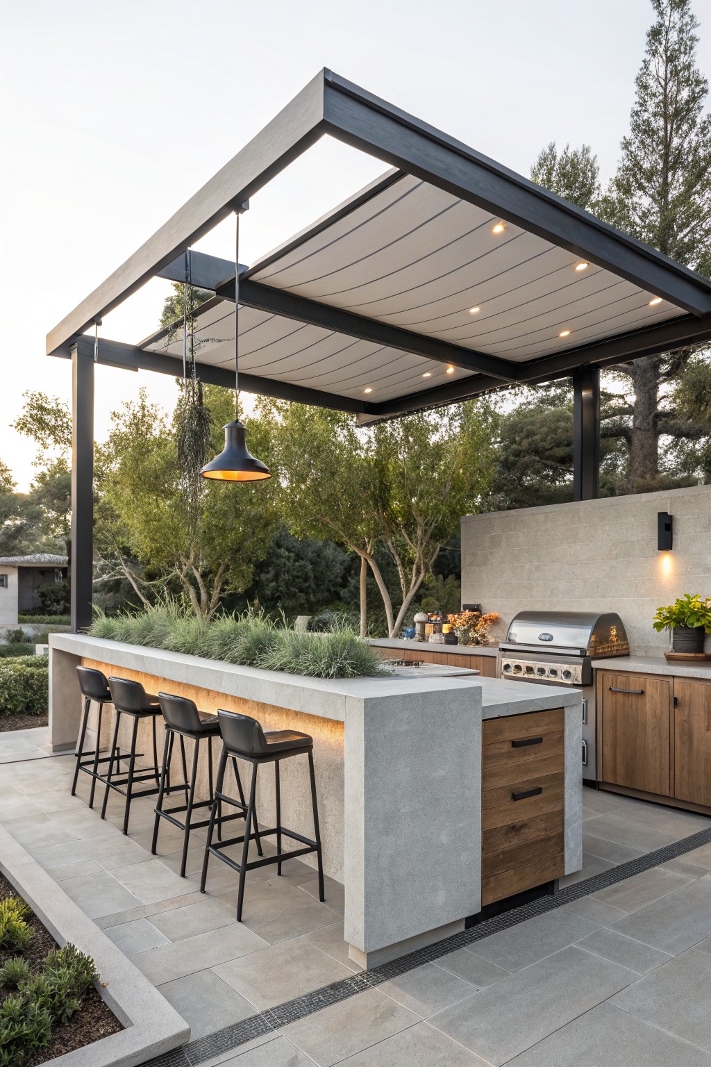 Outdoor kitchen bar with concrete countertop, wood cabinets, stainless steel grill, and four black bar stools under a black steel pergola with white fabric shade and pendant light, surrounded by plants and trees on a gray paver patio.