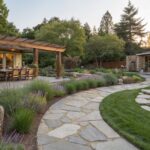 Backyard view showing a house patio with wooden dining table and chairs, nearby lounge seats, a curving flagstone path bordered by lavender plants and grasses leading to a small pond with rocks, edged by lawn and trees in evening light.