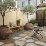Stone-paved courtyard patio featuring a small round metal table with two folding chairs, surrounded by potted plants, olive trees, lavender, a watering can, boxwoods, and a green double door on a beige stucco house wall with stone accents.