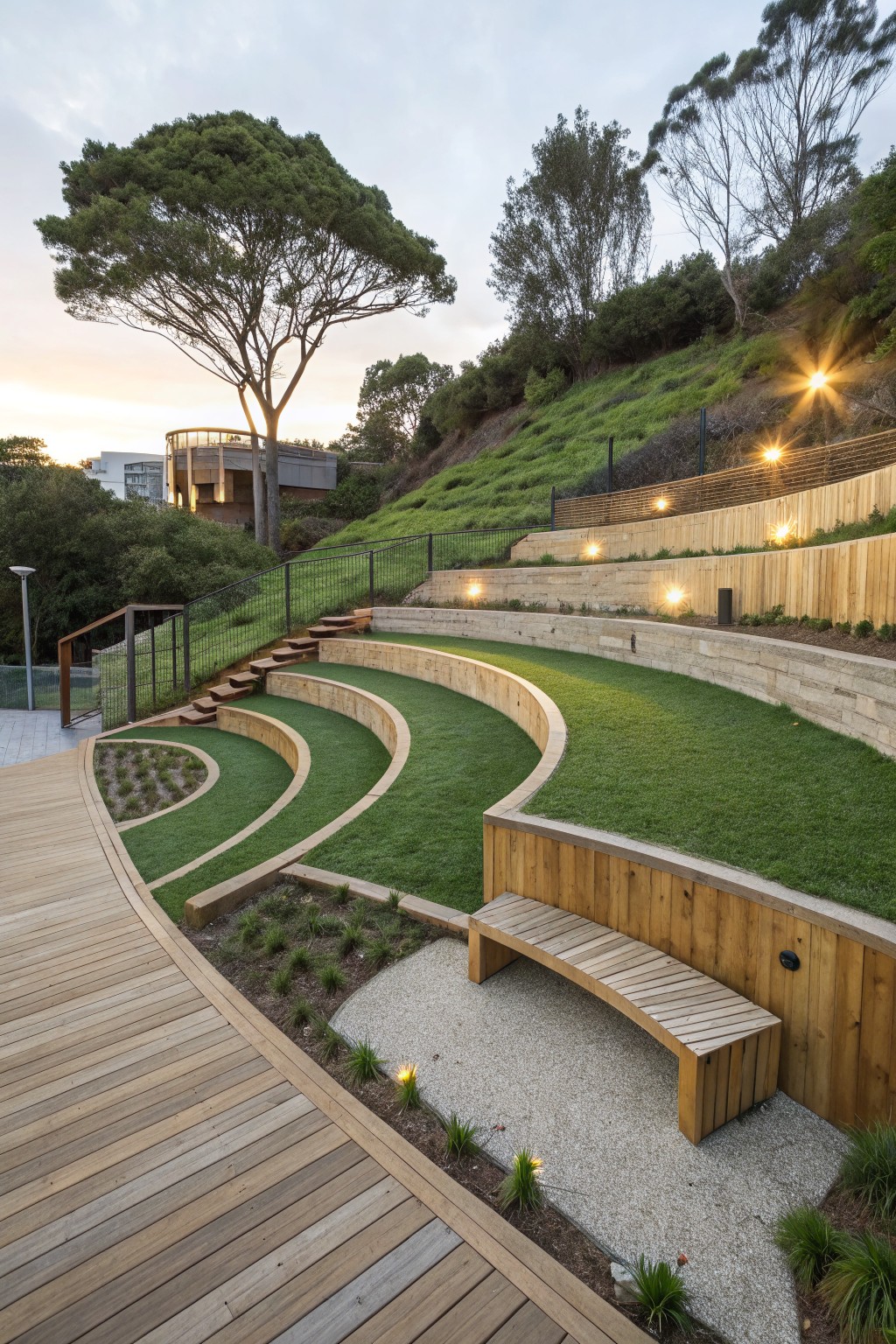 Curved timber-retained terraced steps covered in green grass on a sloped backyard, with wooden pathways, bench, lighting, and surrounding trees and plants at sunset.