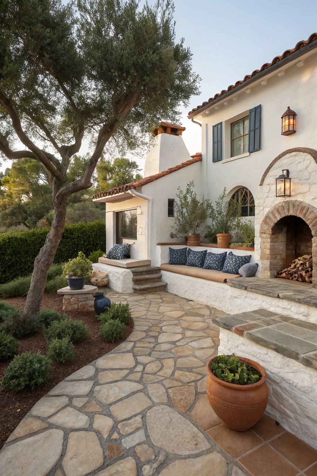 White stucco house wall with red tile roof, arched stone outdoor fireplace stacked with logs, built-in beige bench with blue patterned cushions, stone pathway curving through landscaped area with olive trees, shrubs, and terracotta pots.