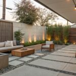 Backyard patio at dusk with rectangular concrete fire pit, adjacent bench and two armchairs, large concrete pavers separated by gravel, low plants, wooden fence, and string lights.