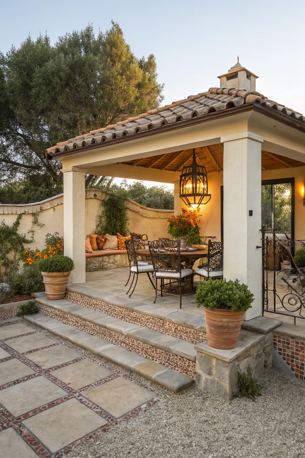 Outdoor gazebo pavilion with terracotta tile roof, round wrought-iron dining table and chairs, built-in cushioned bench along stucco wall, potted plants, lantern light, and stone steps leading up from gravel path.