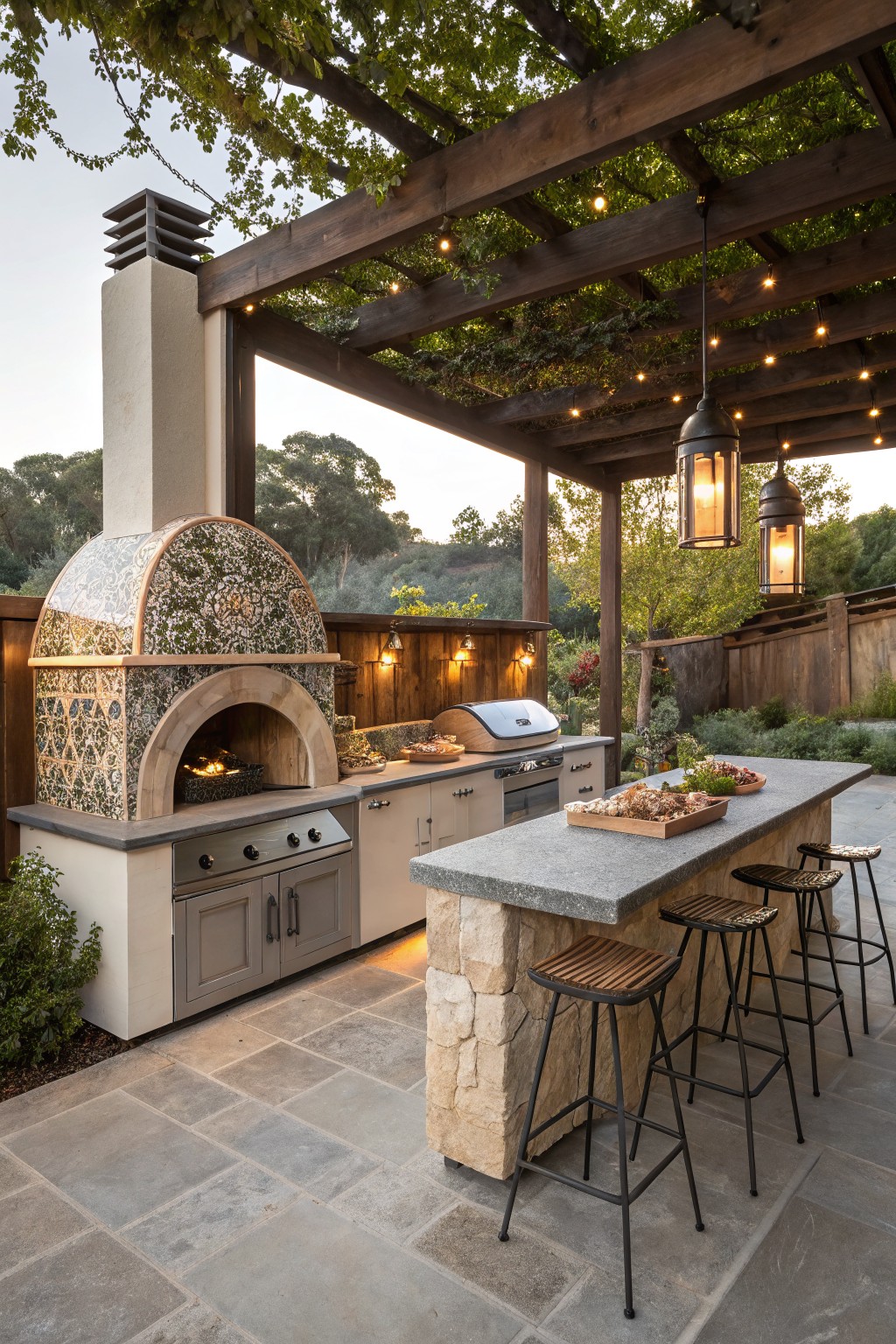 Backyard outdoor kitchen featuring a domed tiled pizza oven, gas grill, gray cabinets, concrete countertop island with wood and metal bar stools, under a vine-covered wooden pergola with hanging lanterns and string lights, on a gray stone patio surrounded by plants and fencing.