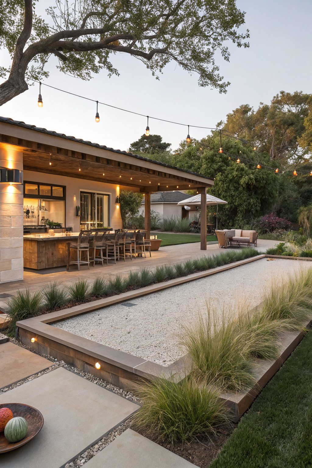 Covered outdoor kitchen with bar stools and wood counter on a patio adjacent to a rectangular bocce court filled with light gravel and edged by grasses, string lights overhead, lounge seating nearby, and trees in the background.